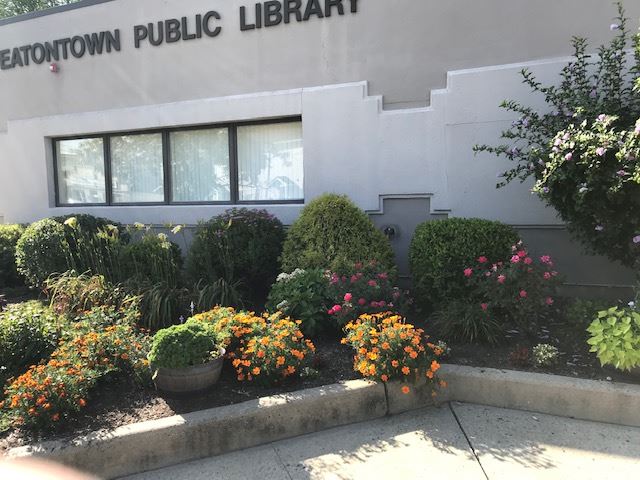Public library exterior sign, white building facade, and landscaped beds with colorful flowers and shrubs.